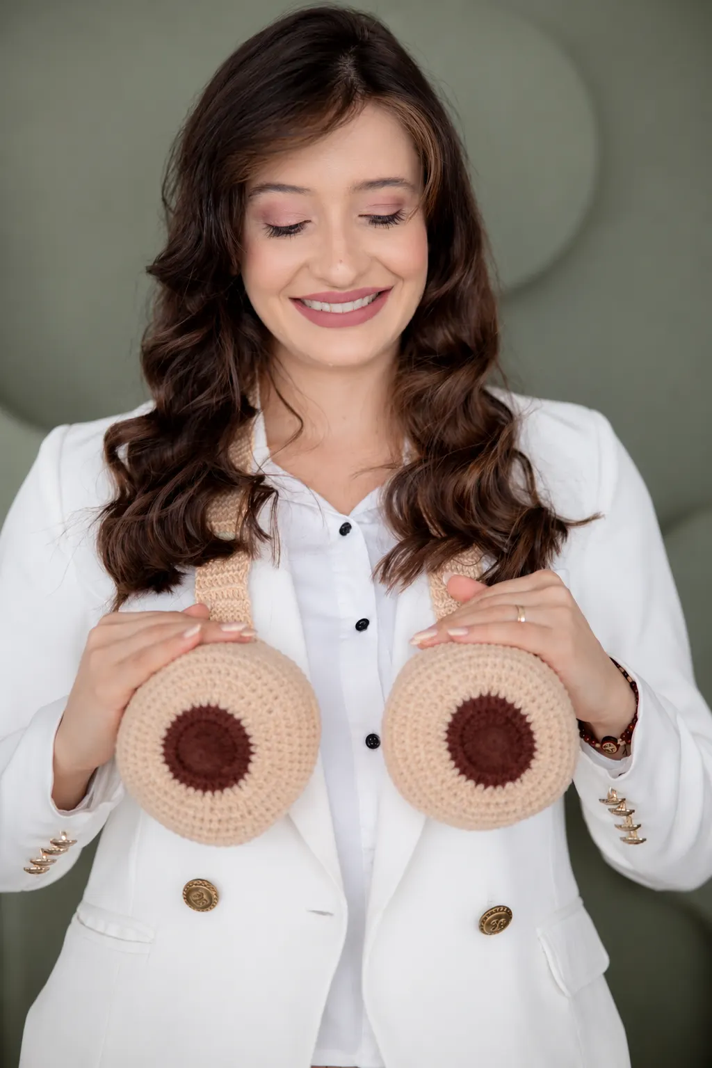 Thaissa Vargas de blazer branco segurando dois modelos de mama em crochê
